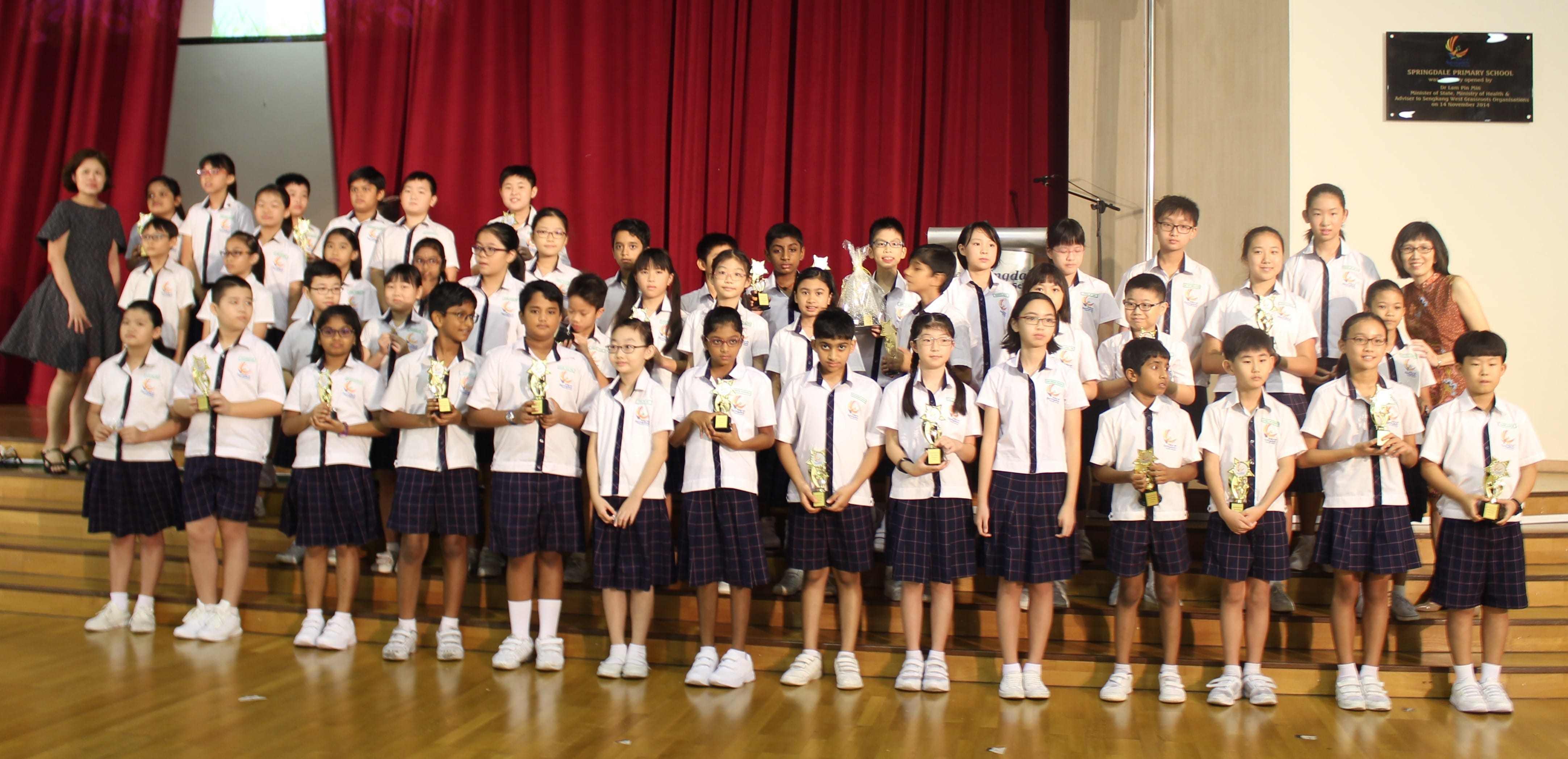 A large group of school children in white uniforms holding trophies posed on a stage with red curtains.